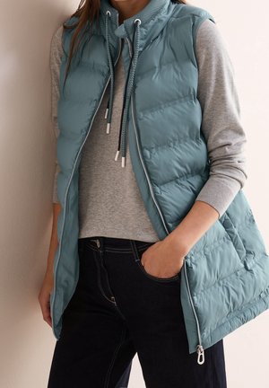 Femme portant une veste matelassée bleue par-dessus un sweat à capuche gris et un jean noir, avec une main dans la poche, debout contre un mur clair.
