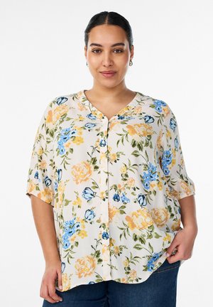 Žena s tmavými vlasmi v kvetovanej košeľovej blúzke so žltými, modrými a zelenými kvetmi stojí na bielom pozadí.