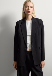 Blazer nero su misura con risvolti appuntiti, chiusura a un bottone, tasche frontali e texture liscia, abbinato a un top bianco a costine.