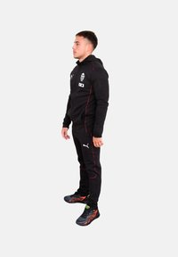 Chándal deportivo negro con detalles rojos, que incluye una sudadera con cremallera y pantalones. Logotipo de la marca en el pecho. Material ligero.