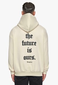 Cremfarbene Kapuzenjacke mit einem großen schwarzen Textdruck auf der Rückseite: "Die Zukunft gehört uns." Das Design umfasst eine Kapuze mit Kordelzug und ribbelte Bündchen.