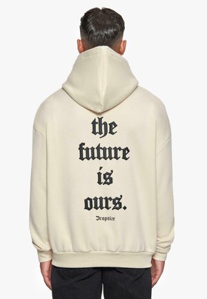 Cremfarbene Kapuzenjacke mit einem großen schwarzen Textdruck auf der Rückseite: "Die Zukunft gehört uns." Das Design umfasst eine Kapuze mit Kordelzug und ribbelte Bündchen.