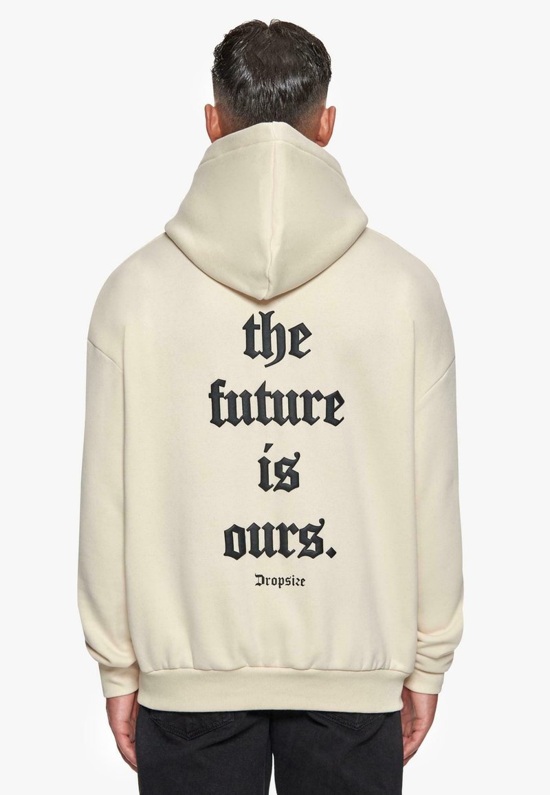 Cremfarbene Kapuzenjacke mit einem großen schwarzen Textdruck auf der Rückseite: "Die Zukunft gehört uns." Das Design umfasst eine Kapuze mit Kordelzug und ribbelte Bündchen.