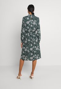 Robe fleurie verte avec manches longues, taille élastique et détail dos avec ouverture en forme de goutte. Motifs floraux blancs sur un tissu texturé. Talons apparents.