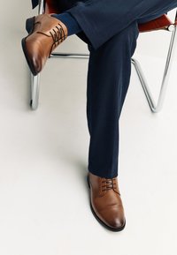 Homme assis sur une chaise, jambes croisées, portant un pantalon foncé et des chaussures habillées en cuir marron poli sur un sol blanc.