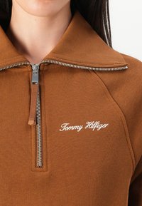 Brown zip-up sweatshirt with a ribbed texture, folded collar, and white "Tommy Hilfiger" logo on the left side of the chest.