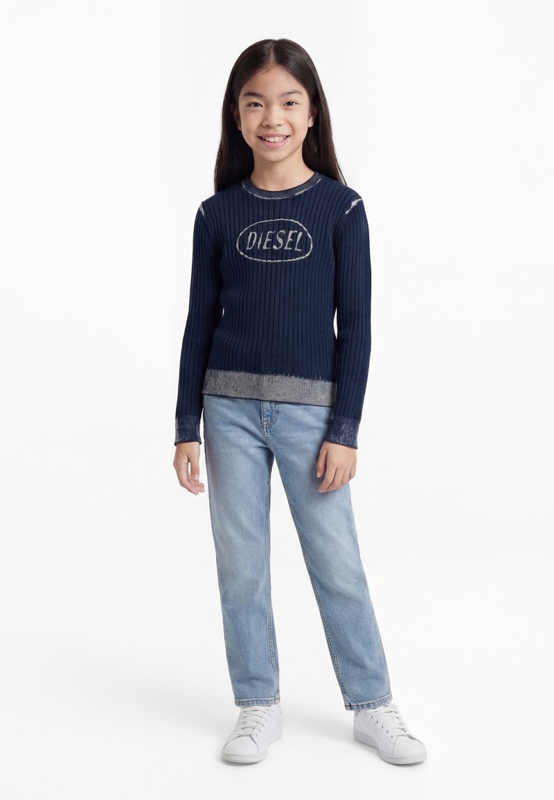 Ung pige smilende, iført en marineblå ribstrikket sweater med "DIESEL" logo, lyseblå jeans og hvide sneakers, stående mod en hvid baggrund.