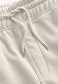 Beige sweatpants med struktureret stof, elastisk talje, og en hvid snor med metaltips, der fremhæver en frontlomme.