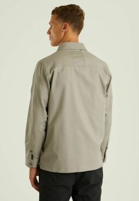 Šviesiai pilkas ilgomis rankovėmis marškinis su apykakle, užpakaline siūle, mygtukais užsegamomis rankovėmis ir lygaus tekstūros audiniu. Dizainas yra paprastas ir funkcionalus.