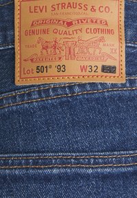 Étiquette des jeans Levi's 501®, en cuir beige avec texte rouge, incluant Lot 501® '93 et taille W32. Fond en denim bleu, coutures visibles.