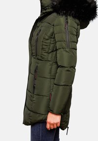 Vue de côté d'une personne portant une veste d'hiver matelassée vert foncé avec des poches zippées et une capuche bordée de fourrure noire, associée à un jean bleu.