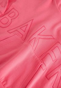 Vaaleanpunainen collegepaita, jossa on kohokuvioitu "BAKER" -teksti, kengurupussi ja ribattu hihansuut. Pehmeä materiaali ja rento muotoilu.