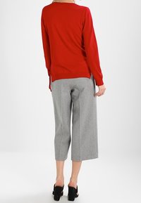 Maglione rosso a maglia con scollatura rotonda e lacci laterali, abbinato a pantaloni leggeri grigi a gamba larga. Tacchi neri open-toe completano il look.