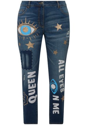 Denim jeans i mørkeblå har glitrende guldstjerner og tekstgrafik. Slidte lapper og øjemotiver tilføjer visuel interesse og tekstur.