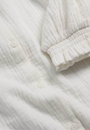 Puha, fehér, texturált szövet, összegyűrt elasztikus mandzsettával és apró fehér gombokkal egy könnyű hosszú ujjú ruhadarabon.