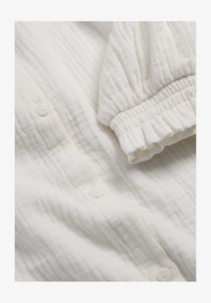 Mekani bijeli teksturirani materijal s naboranim elastičnim manšetama i malim bijelim gumbima na laganom dugom rukavu.