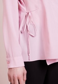 Lichtroze blouse met lange mouwen en details van een zijstrik, met geknoopte manchetten, gedragen met zwarte broek, hand ontspannen aan de zijde.