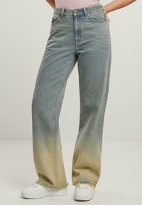 Jeans de mezclilla azul claro con un lavado desgastado, diseño de pierna ancha, dobladillos deshilachados y estilo de cinco bolsillos. Combinados con zapatillas blancas.