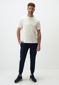 Heren crème short-sleeve t-shirt gecombineerd met marineblauwe tapered broeken en zwarte sneakers. Glad materiaal, ontspannen pasvorm, subtiel ontwerp.
