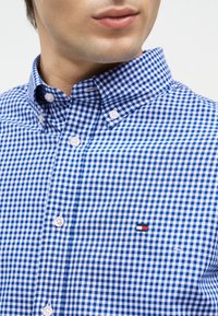 Κοντινό πλάνο ενός άνδρα που φοράει ένα μπλε και λευκό καρό πουκάμισο με κουμπιά και ένα μικρό ορθογώνιο λογότυπο στο στήθος.