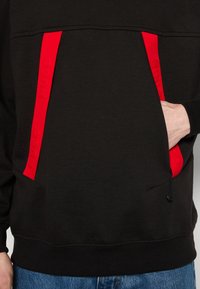 Felpa nera con dettagli rossi sulle tasche anteriori. Presenta una tasca a marsupio foderata e una vita elastica, realizzata in tessuto morbido.