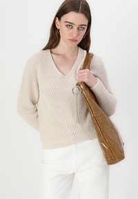 Pull en tricot beige avec un col en V, présentant un motif texturé ; associé à un sac à épaule marron et brillant. Pantalons blancs.