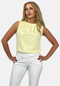 Bluzka bez rękawów w miękkim żółtym kolorze, z zakładkami przy dekolcie. W duecie z białymi dopasowanymi spodniami, tworząc zrównoważoną sylwetkę.