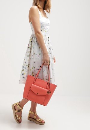 Korallenfarbene Ledertasche mit einer Vorderstasche, zwei Griffen und einem runden Logo-Anhänger; kombiniert mit Keilsandalen mit goldenen Riemen und einem Blumenkleid.