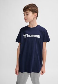 Tricou din bumbac bleu marin cu mâneci scurte, având un mare logo alb "hummel" pe piept. Purtați cu pantaloni gri deschis.