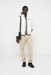 Witte gewatteerde jas met golvend patroon, zwarte rits, verstelbare capuchon. Bruine trui eronder, beige broek en zwarte schoenen maken de outfit compleet.