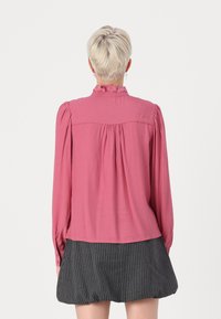 Long-sleeve, pink blouse with a ruffled collar and pleated back. Soft fabric, fitted wrists, and a relaxed fit. Paired with a striped skirt.