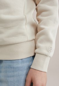 Bež sweatshirt z ribanim manšetom in robom, gladka tekstura z lahkimi šivi in majhnim embosiranim logotipom na rokavu.