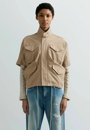 Persona che indossa una giacca beige a maniche corte con tasche a patta sopra una maglia a maniche lunghe e jeans blu strappati con coulisse anteriori.