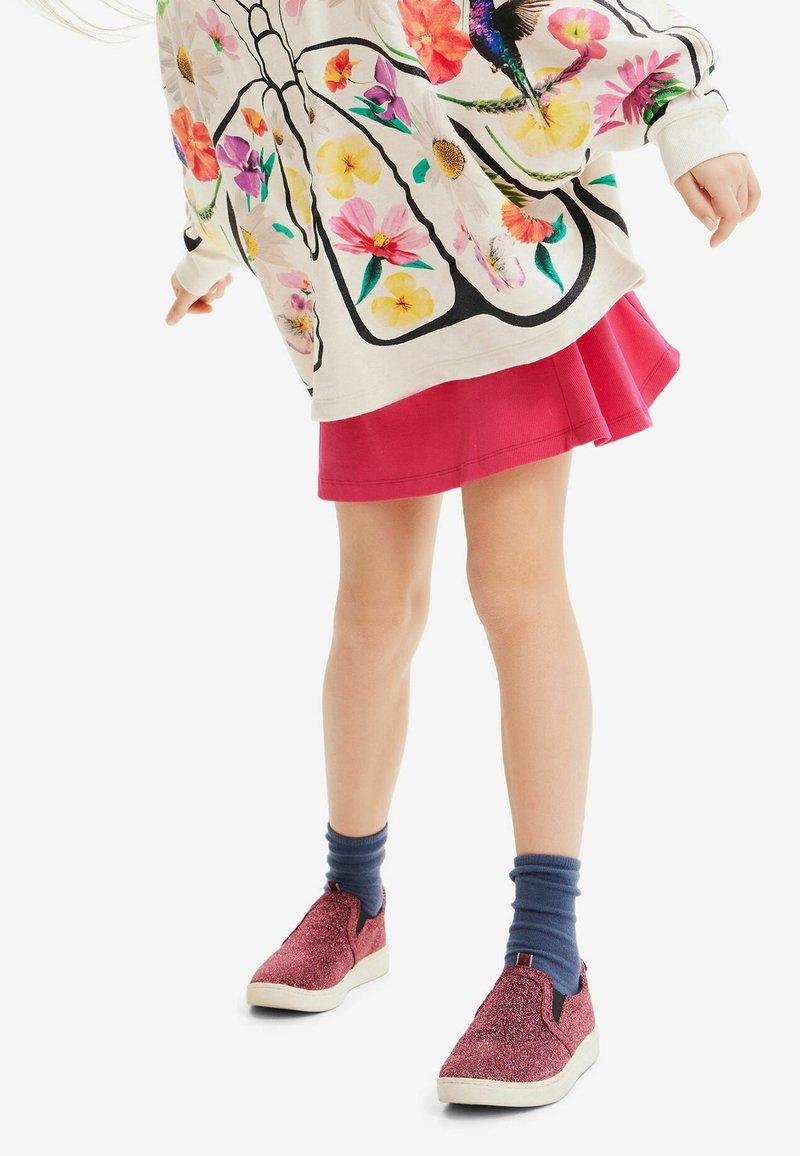 Světlý oversized svetr s barevným květinovým vzorem, doplněný červenou skládací sukní a červenými třpytivými slip-on teniskami. Viditelné modré ponožky.