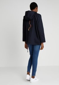 Person som bär en marinblå huvtröja med ankare och kedja på ryggen, blå skinny jeans och vita sneakers, står mot en enfärgad bakgrund.