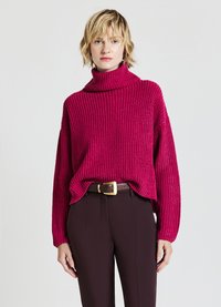 Fuchsia Ripp-Turtleneck-Pullover mit lockerer Passform, langen Ärmeln und subtiler Textur, kombiniert mit hochgeschnittenen braunen Hosen und einem Gürtel.