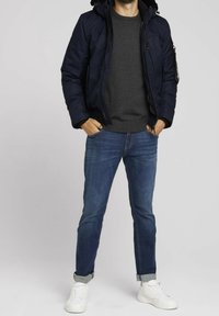 Giacca puffer di un blu navy scuro con cappuccio, materiale leggero, chiusura frontale con zip, accenti neri. Indossata con un maglione grigio e jeans blu. Sneakers bianche.