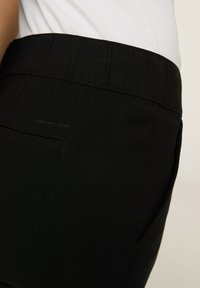 Czarne spodnie o gładkiej fakturze i elastycznym pasku w talii. Posiadają tylną kieszeń oraz subtelny akcent logo na pasku.