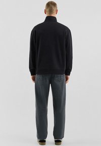 Schwarzer Pullover mit hohem Kragen, aus strukturiertem Stoff. Kombiniert mit locker sitzenden dunkelgrauen Jeans und schwarzen Sneakern.