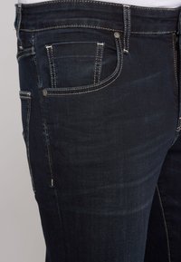 Dunkelblaue Jeans mit Slim-Fit, gesteppten Taschen und kontrastierenden, hellen Nähdetails. Weiche Textur mit leichten Verwaschungen.