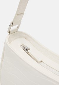 Zbliżenie na białą torebkę z faux skóry krokodyla, ukazujące srebrny zamek błyskawiczny, mocowanie paska oraz fakturę powierzchni.