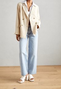 Beżowa marynarka dwurzędowa z złotymi guzikami, w połączeniu z jasnoniebieskimi spodniami typu wide-leg i białymi sandałami na platformie. Minimalistyczny design, luźny krój.