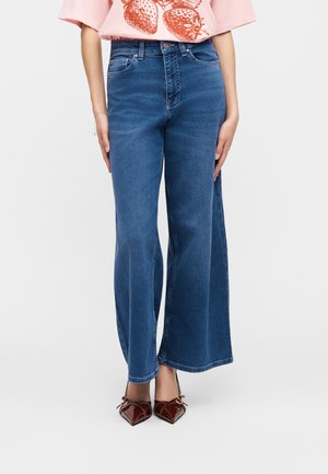 Woman wearing high-waisted blue wide-leg jeans, pink t-shirt with strawberry print, and brown pointed-toe heels with bows.