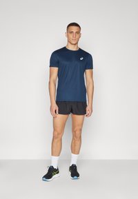 Tumed sinine spordipe shirt, lühikeste varrukatega, väikese logoga. Kombineeritud mustade jooksutossudega ja mustade spordishortsidega, millel on rohelised ja sinised detailid.