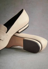 Mocassins en cuir beige avec un bout arrondi, un petit talon carré et une semelle en caoutchouc noire ; lignes épurées et détails minimalistes pour un look moderne.