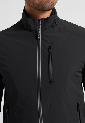 Man wearing black zip-up jacket with chest and side zippered pockets, high collar, and "Milestone" logo on front zipper pull.