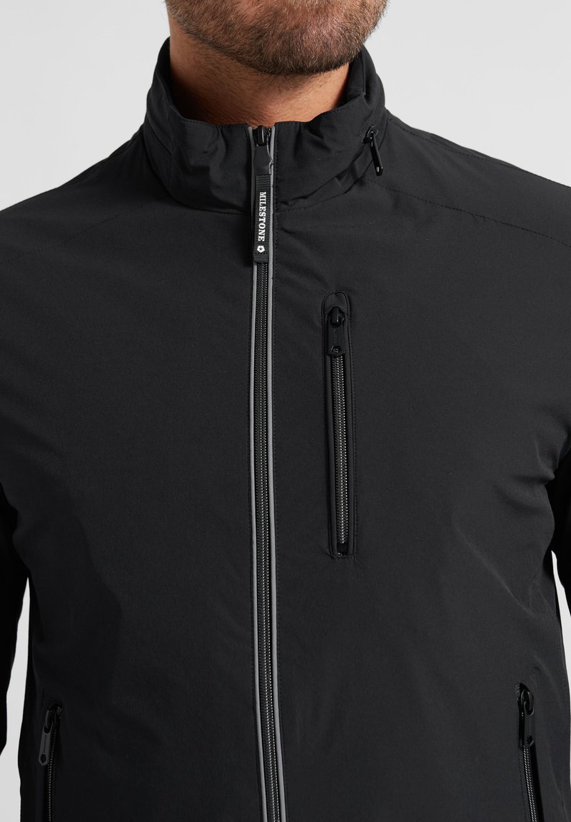 Mann trägt schwarze Reißverschlussjacke mit Brust- und Seitentaschen, hohem Kragen und "Milestone"-Logo am vorderen Reißverschlussanhänger.