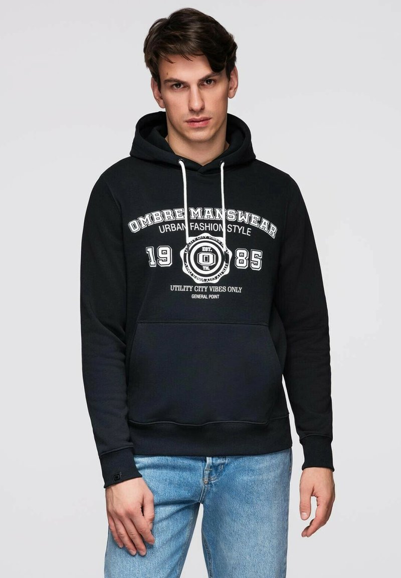 Sweat à capuche noir avec texte graphique blanc et design, comprenant une poche avant, une capuche avec cordon de serrage et des poignets côtelés aux manches.