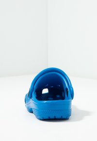 Modré gumové kloboukové boty s perforací a pásem na patě, pohled z boku na bílé ploše a pozadí rohu zdi.