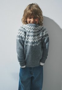 Maglione grigio a maglia con un motivo a zigzag bianco sullo scollo, abbinato a pantaloni in denim blu. Il maglione presenta polsini e orlo a costine.